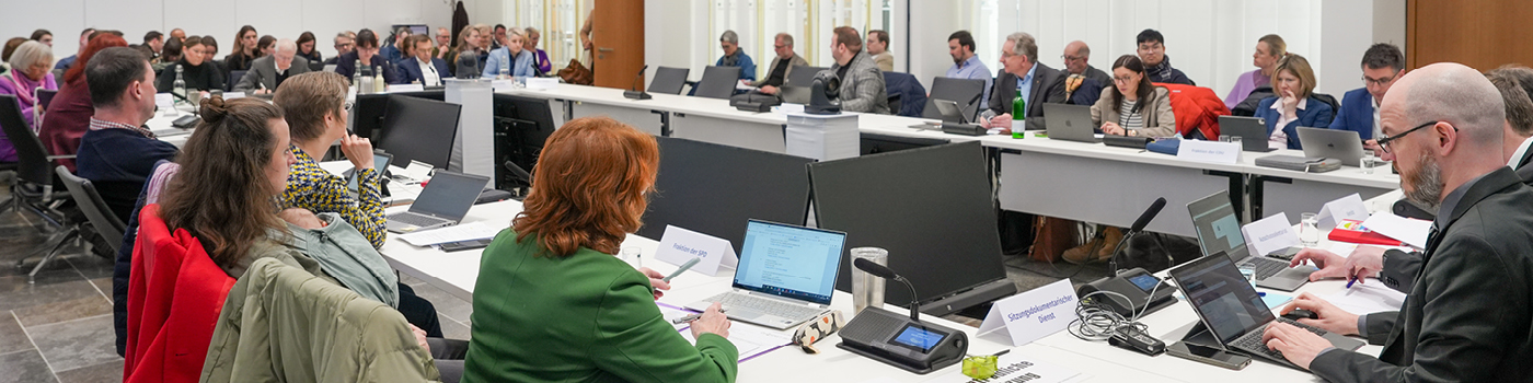 Zu sehen ist ein gro&szlig;er Konferenzraum, in dem viele Menschen an U-f&ouml;rmig angeordneten Tischen sitzen. Die Teilnehmenden haben Laptops, Unterlagen und Mikrofone vor sich.